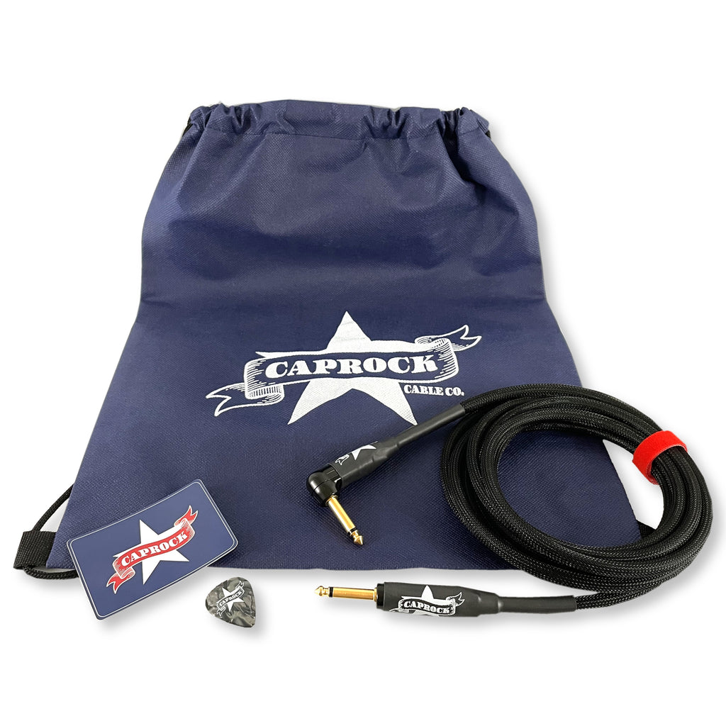 A Caprock Cable Co. product set on a white background, featuring a navy blue drawstring bag with a white star logo, a coiled black audio cable with gold-toned connectors and a red loop, a branded sticker, and a gray guitar pick.