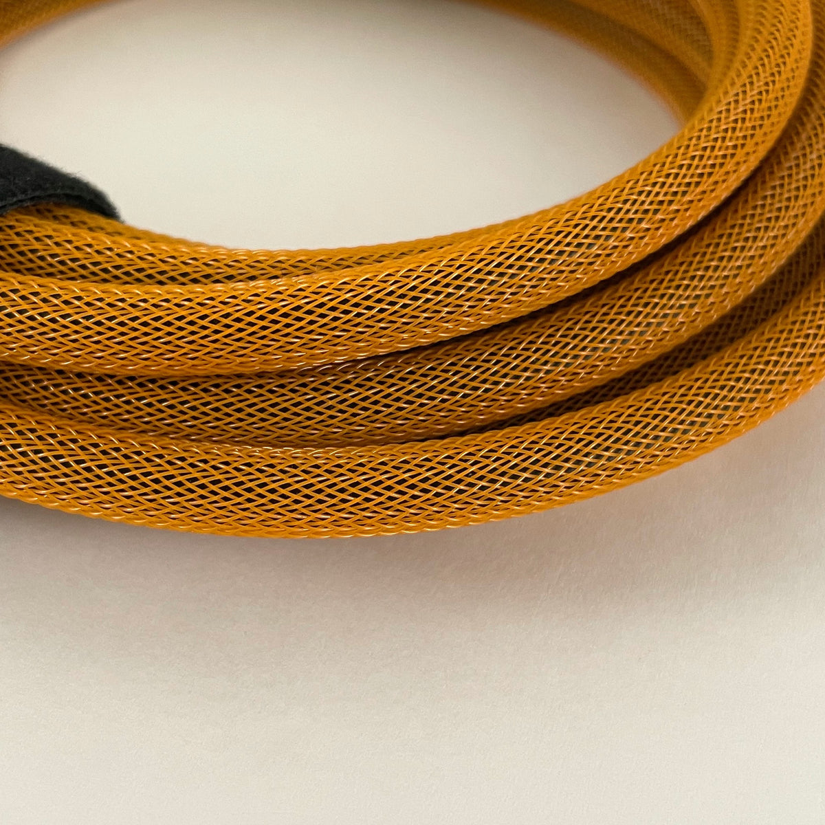 A close-up of a coiled orange braided cable with a fine mesh texture, resting on a plain off-white surface.